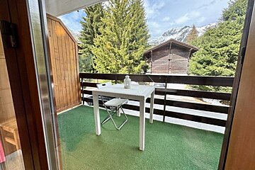 Balcony with a white table and chair on green turf, overlooking a wooden chalet and snow-covered mountains.