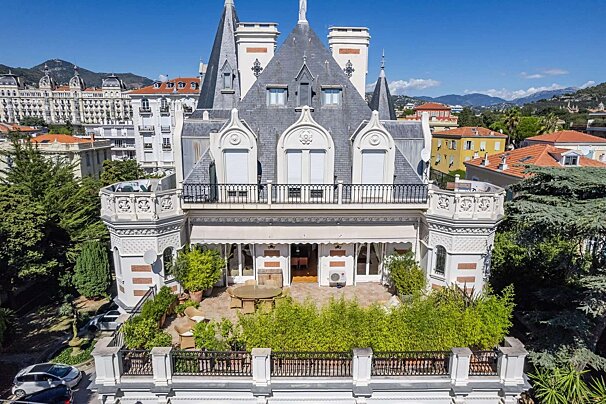 An ornate, multi-spired villa with a large patio surrounded by lush greenery. It stands in a sunny city with mountains in the background.