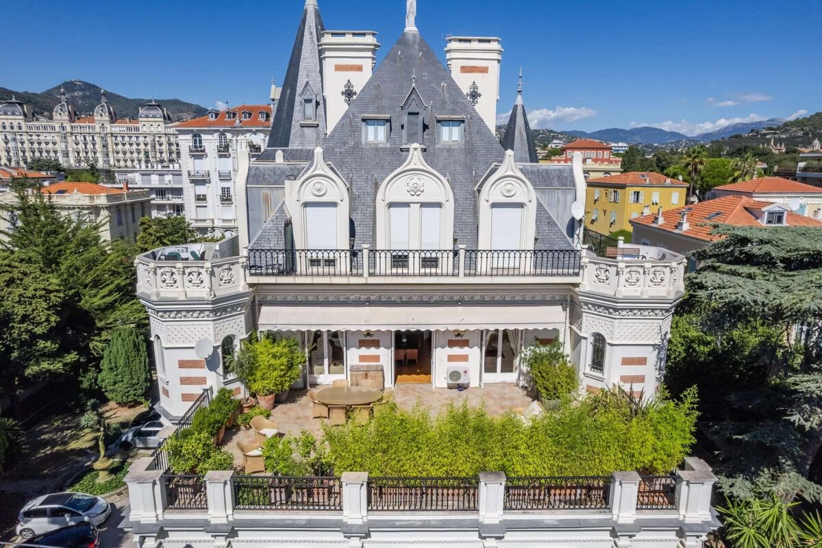 A grand, ornate white mansion with spires and a dark grey roof, featuring balconies, a large patio with plants, and outdoor seating. Mountains and city buildings are in the background.