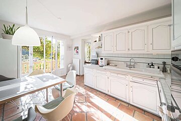 Bright, sunlit kitchen and dining area with white cabinets, terracotta floors, and French doors opening to a scenic balcony overlooking trees.