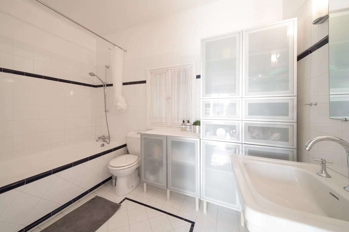 A bright, modern bathroom with white tiles, black trim, bathtub, toilet, sink, and white cabinets with frosted glass doors.