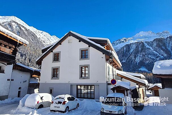 A snowy Alpine village features white buildings with dark roofs, snow-covered cars, and majestic snow-capped mountains under a clear blue sky.