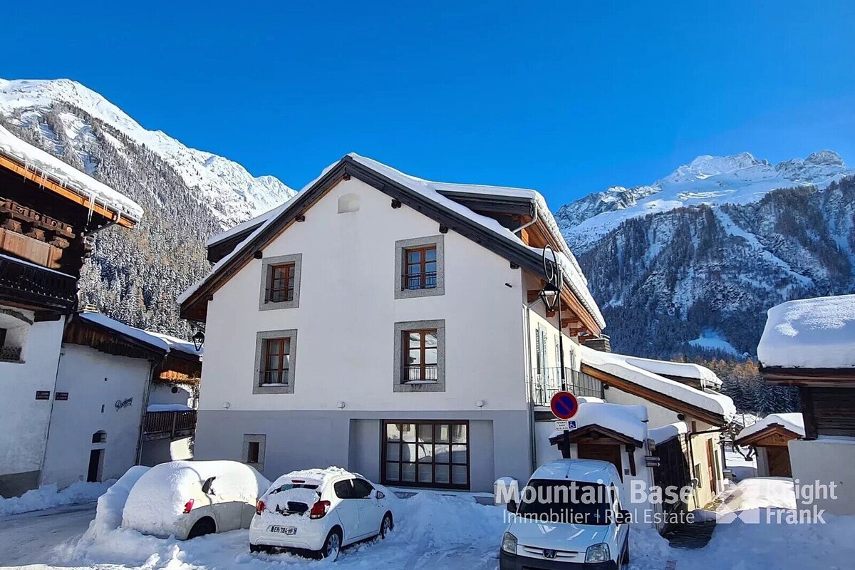 A snowy Alpine village features white buildings with dark roofs, snow-covered cars, and majestic snow-capped mountains under a clear blue sky.