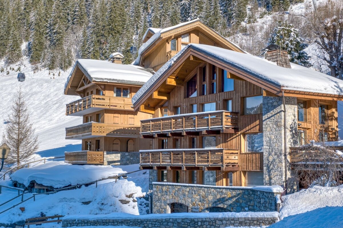 Luxurious wooden and stone chalets with snow-covered roofs and balconies overlook a sunny ski slope and pine forest in a winter mountain resort.
