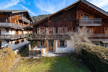 Two traditional wooden chalets with balconies, a green yard, and distant mountains under a blue sky.