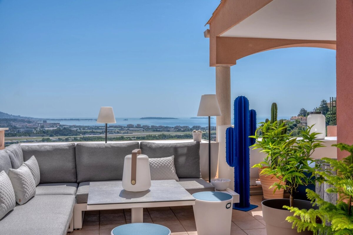 A modern rooftop terrace with a grey sofa, quirky blue cactus, and potted plants, overlooking a panoramic sea and city view under a clear sky.
