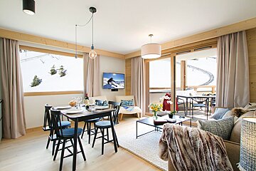 A chic ski chalet living/dining area featuring a set table, cozy sofa, TV, and breathtaking snowy mountain views from windows and balcony.