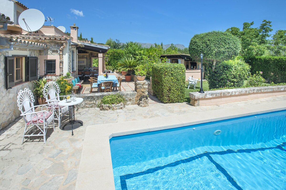 A sunny stone villa with a vibrant blue swimming pool, outdoor patio, and lush green garden under a clear blue sky.