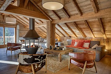 Cozy wooden chalet living room with a modern suspended fireplace, leather chairs, and a mountain view from the balcony.