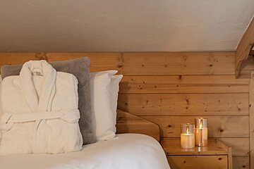 Cozy bedroom with white bathrobe on bed, wooden walls, and lit candles on a nightstand, creating a warm ambiance.