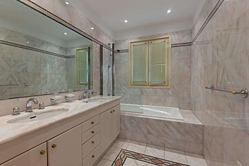 A light-colored tiled bathroom features a double vanity with a large mirror, a built-in bathtub, a window with blinds, and decorative floor tiles.