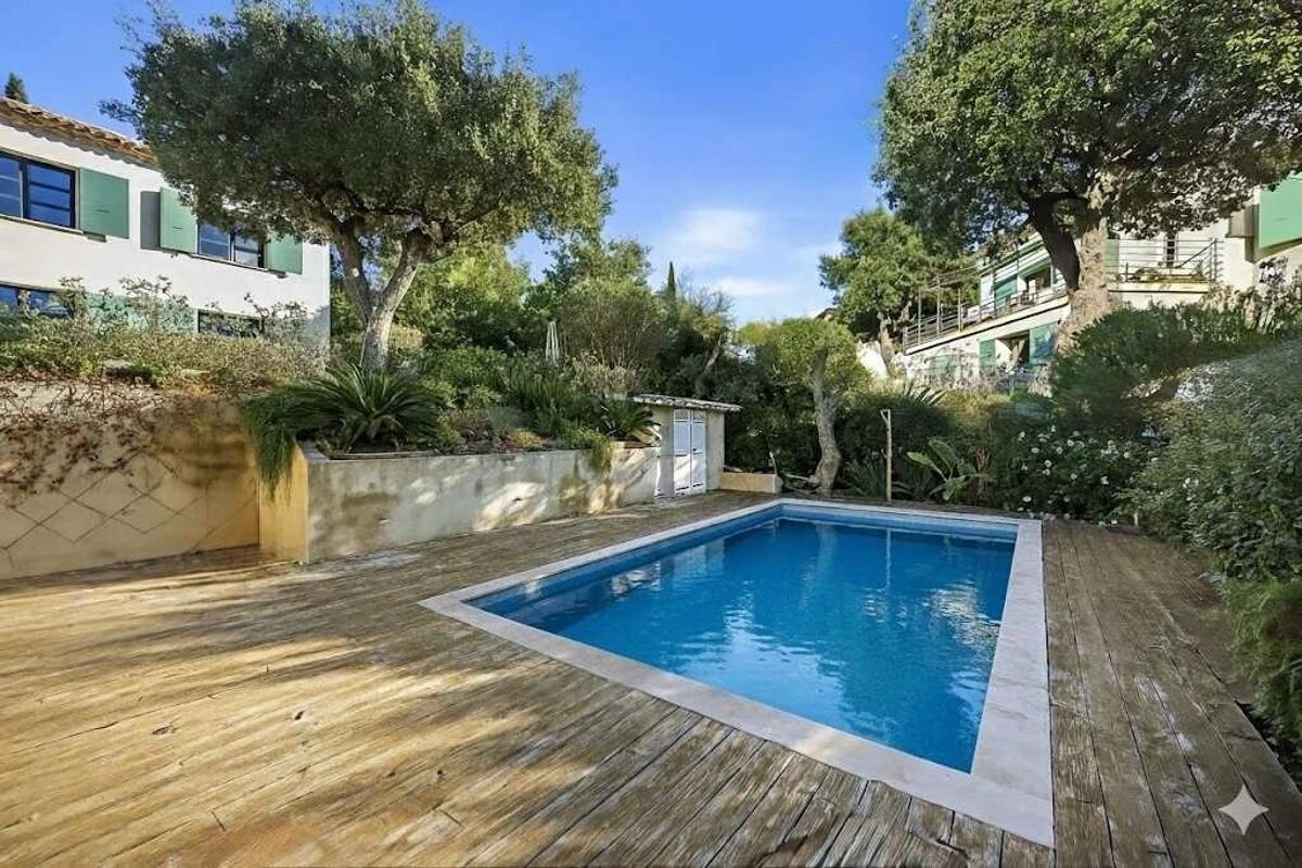 A rectangular swimming pool with a wooden deck, set in a lush garden with trees and buildings under a clear blue sky.