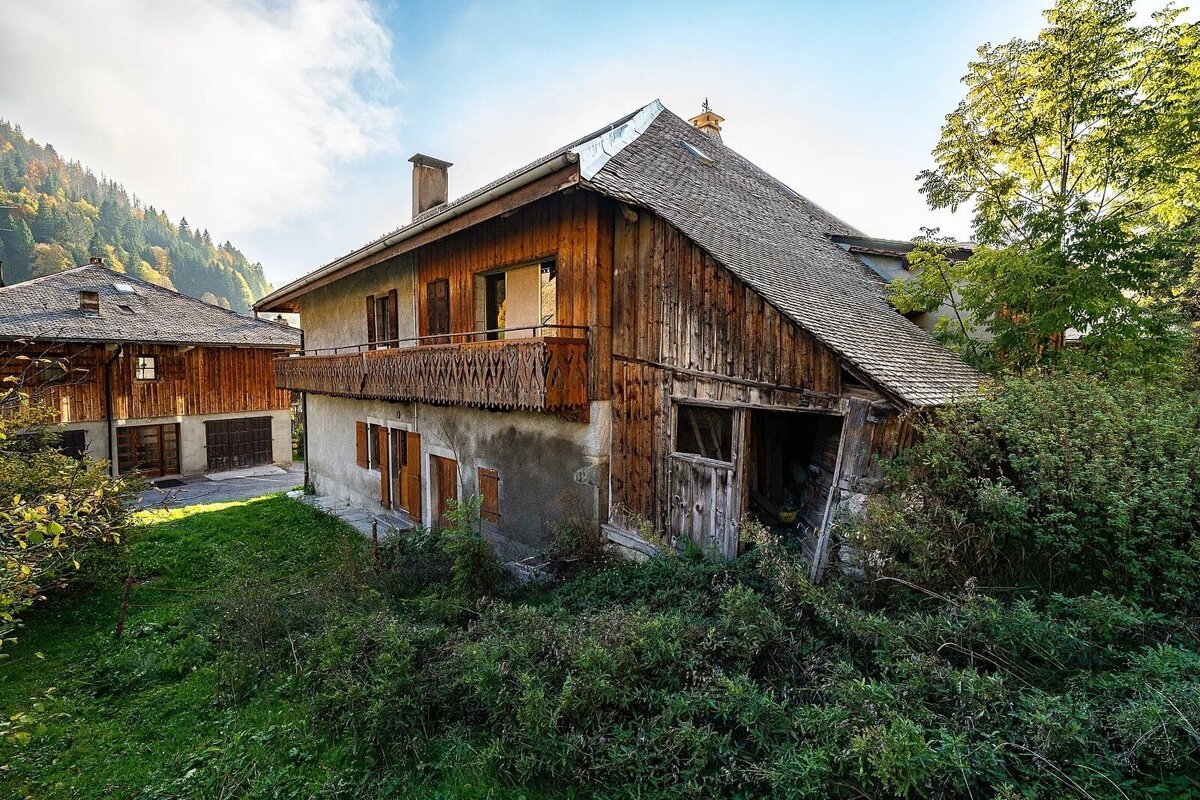 Rustic mountain homes, one with a wooden balcony, surrounded by lush green foliage under a partly cloudy blue sky.