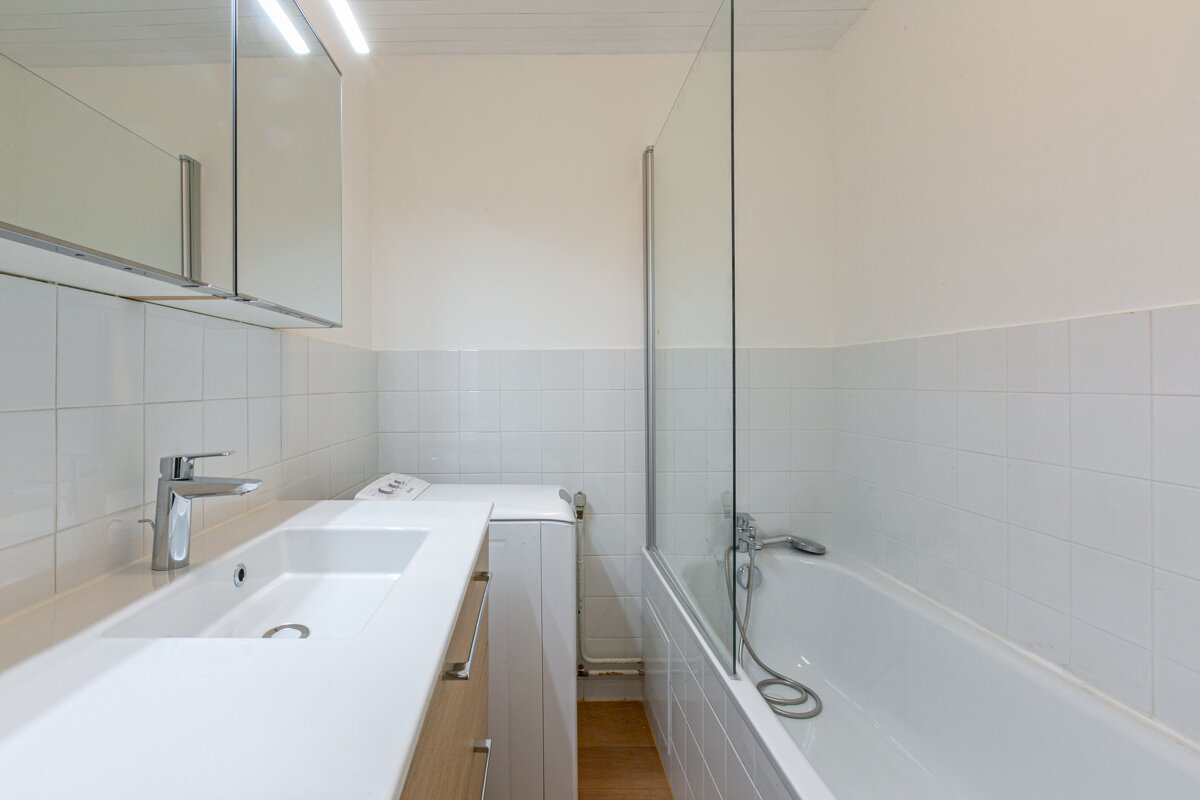 Bright, modern bathroom with a vanity sink, integrated washing machine, and a tiled bathtub with a glass shower screen.
