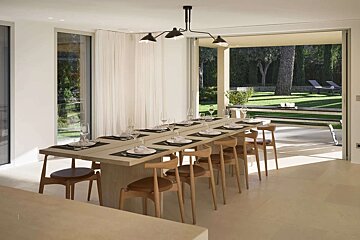 A stylish dining room with a long table, sleek black chandelier, and large windows revealing a beautiful green garden.