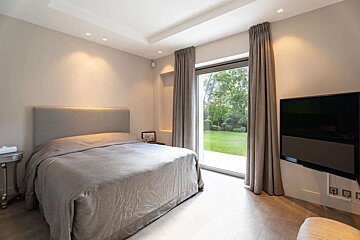 A modern bedroom with a grey bed, wall-mounted TV, and a large glass door opening to a lush green lawn. Recessed lighting illuminates the neutral-toned space.