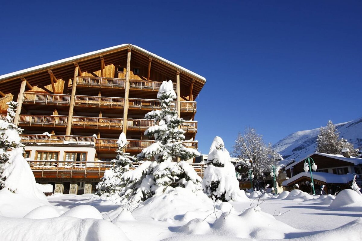 A large wooden chalet stands under a clear blue sky, surrounded by snow-covered pine trees on a sunny winter day.