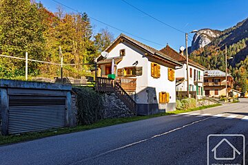 A white house with wooden shutters and a balcony on a sunny autumn day, situated by a road with a garage below. Mountains and colorful trees are in the background.