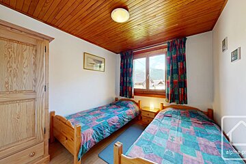 Cozy bedroom featuring two single wooden beds, a wardrobe, a wooden ceiling, and a window with colorful curtains.