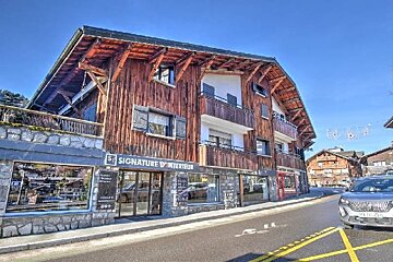 An alpine chalet-style building featuring a ground-floor shop labeled 'Signature d'Interieur' along a road with a parked car.