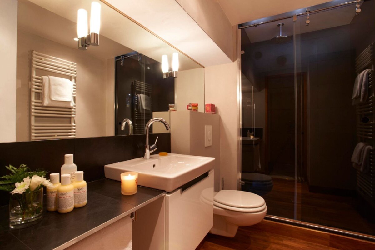 A modern bathroom with a white sink, toilet, and glass shower. Features include a large mirror, towel warmer, toiletries, flowers, and a lit candle.