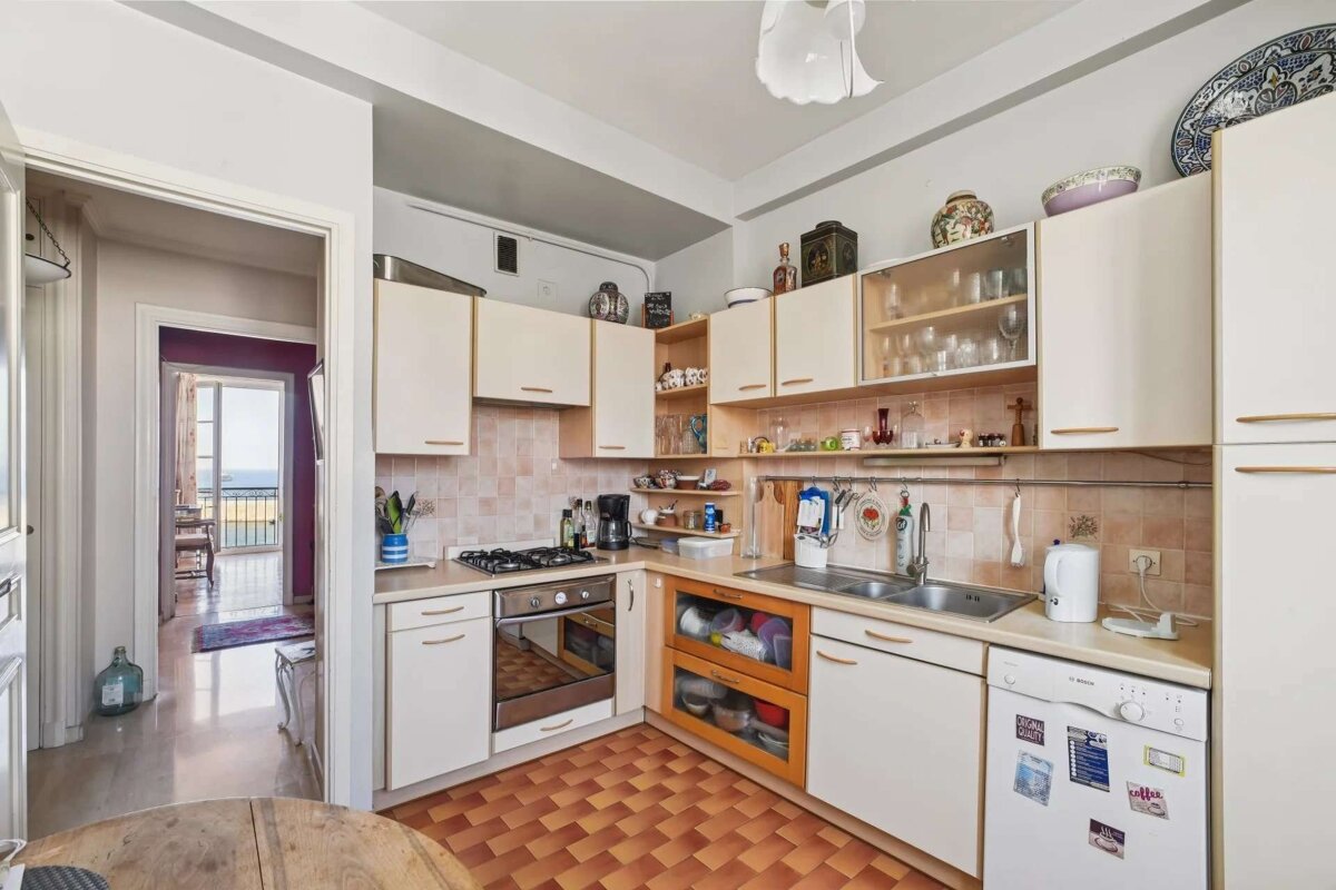 A cozy kitchen with cream cabinets, a tiled backsplash, and a checkered floor. An open doorway reveals another room with a balcony overlooking the sea.