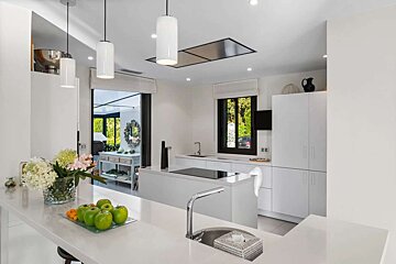 A sleek, modern white kitchen features a large island, pendant lights, and green apples. Windows overlook lush greenery.