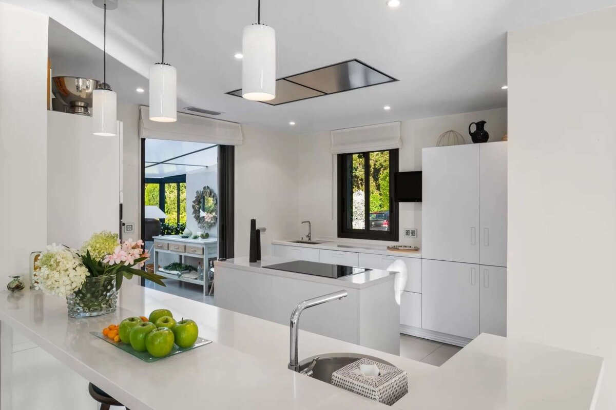A bright, modern all-white kitchen with a central island, sleek appliances, and green apples on the counter, bathed in natural light.