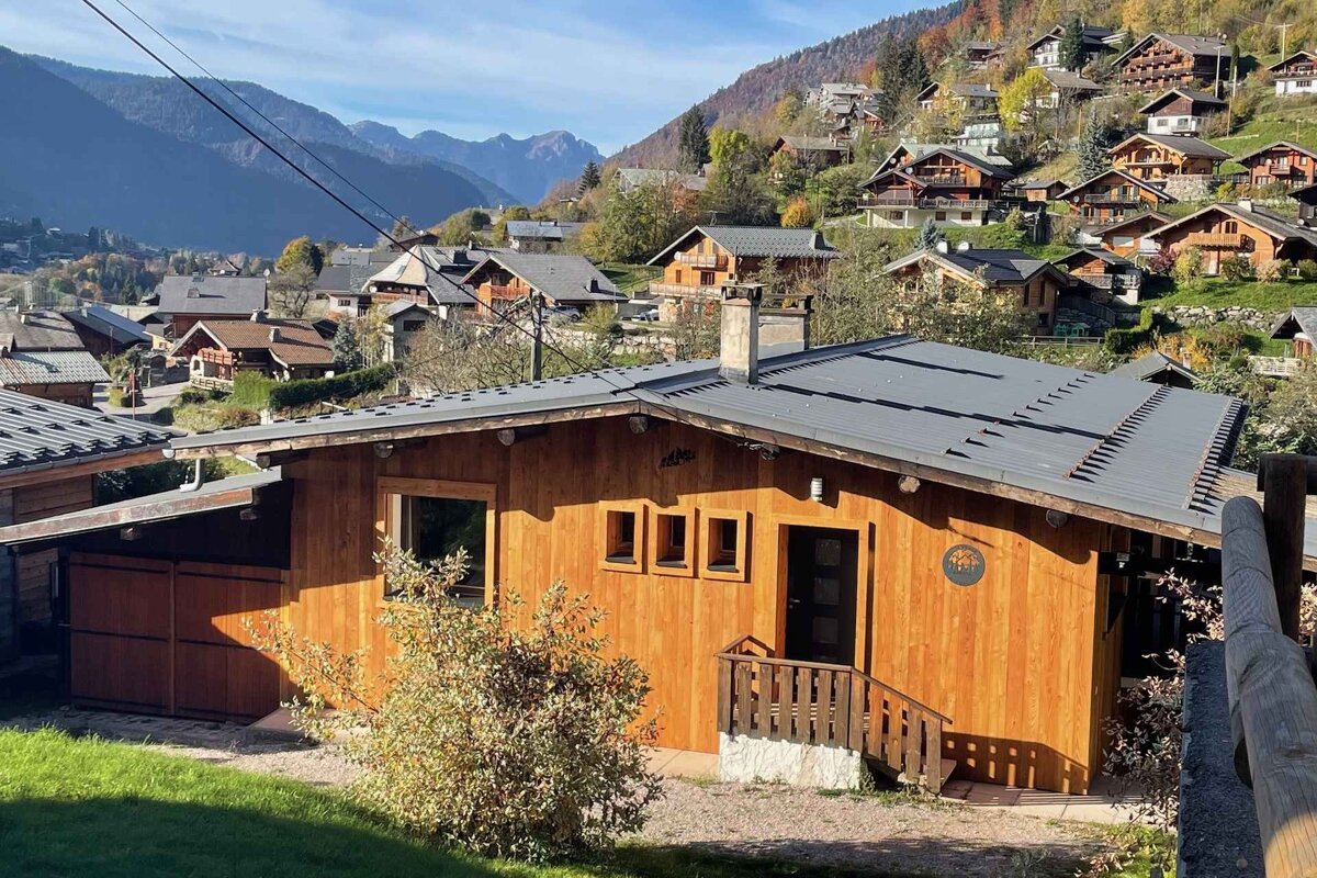 Sunny wooden chalet in a picturesque mountain village, surrounded by other houses on terraced hillsides, under a clear sky.