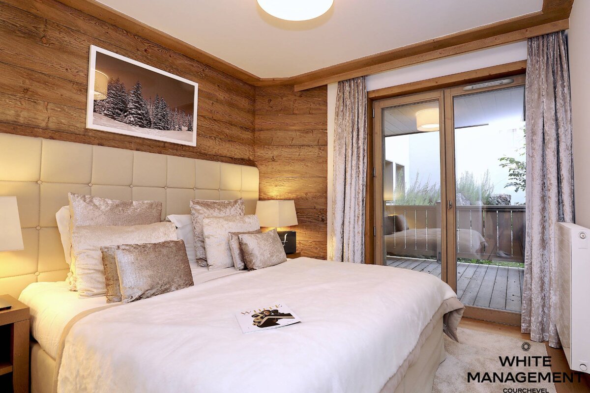 Cozy chalet bedroom with wood paneling, a large bed with neutral-toned pillows, and glass doors opening to a balcony.