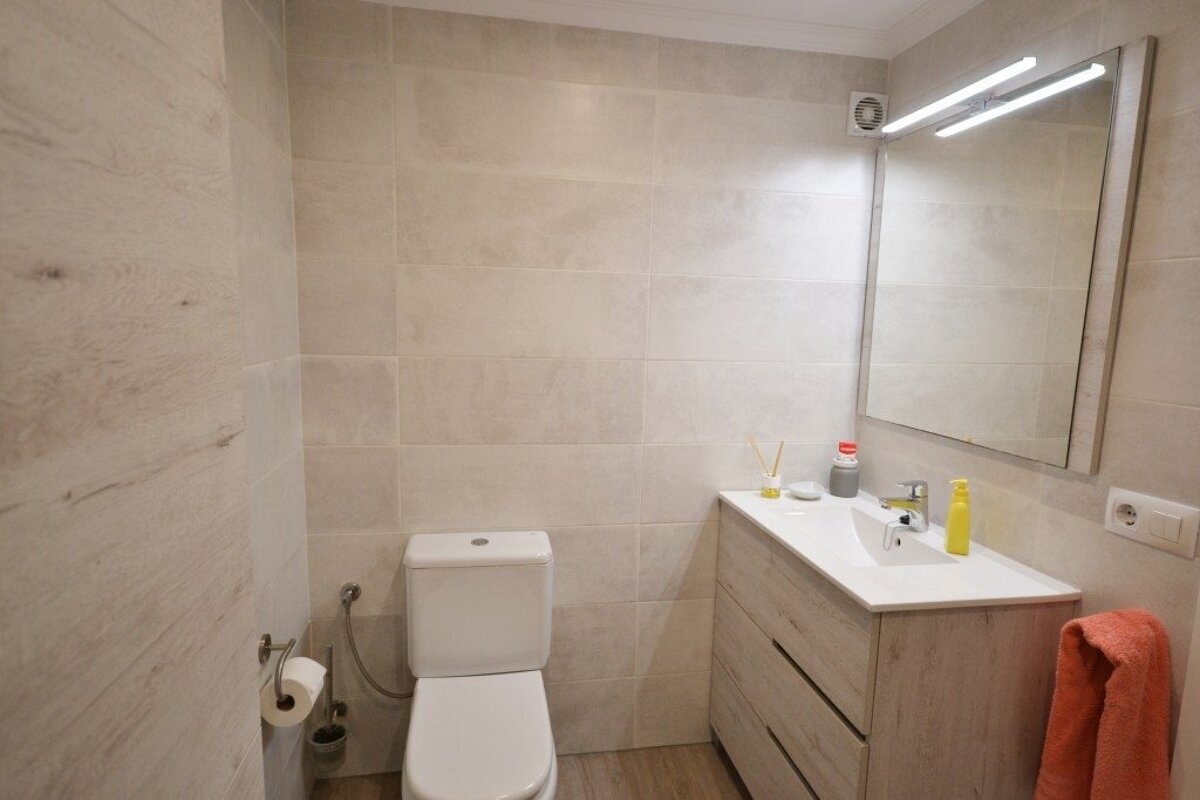 A modern beige-tiled bathroom featuring a white toilet, a light wood vanity cabinet with a sink, and a large illuminated mirror.