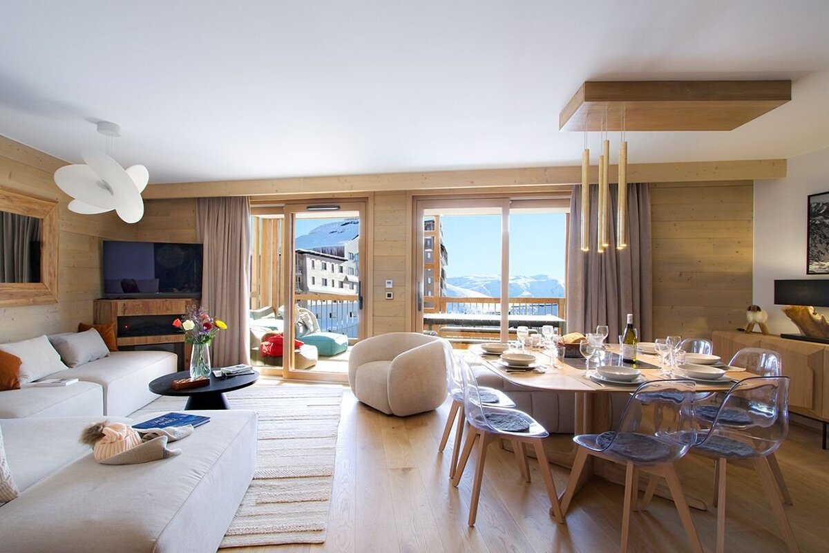 A cozy, modern open-plan living and dining room with wooden accents, large windows offering snowy mountain views, and a set dining table.