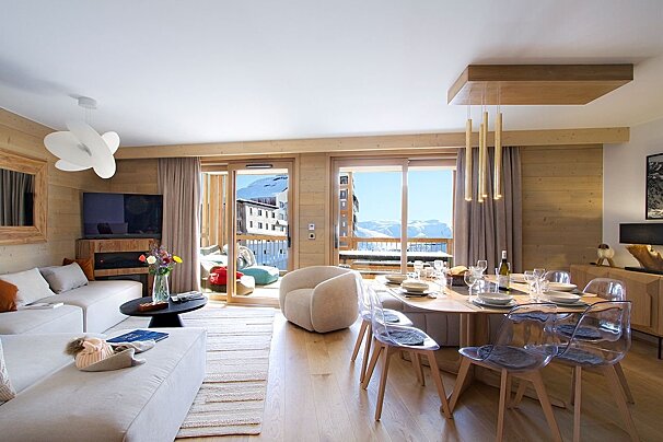 A bright, modern ski apartment living/dining room. Features light wood, white furniture, a set dining table, and large windows revealing snowy mountain views.