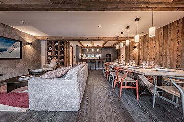 A modern, open-plan chalet interior with a long dining table, grey sofa, and integrated kitchen. Features rustic wood accents, beams, and stylish lighting.