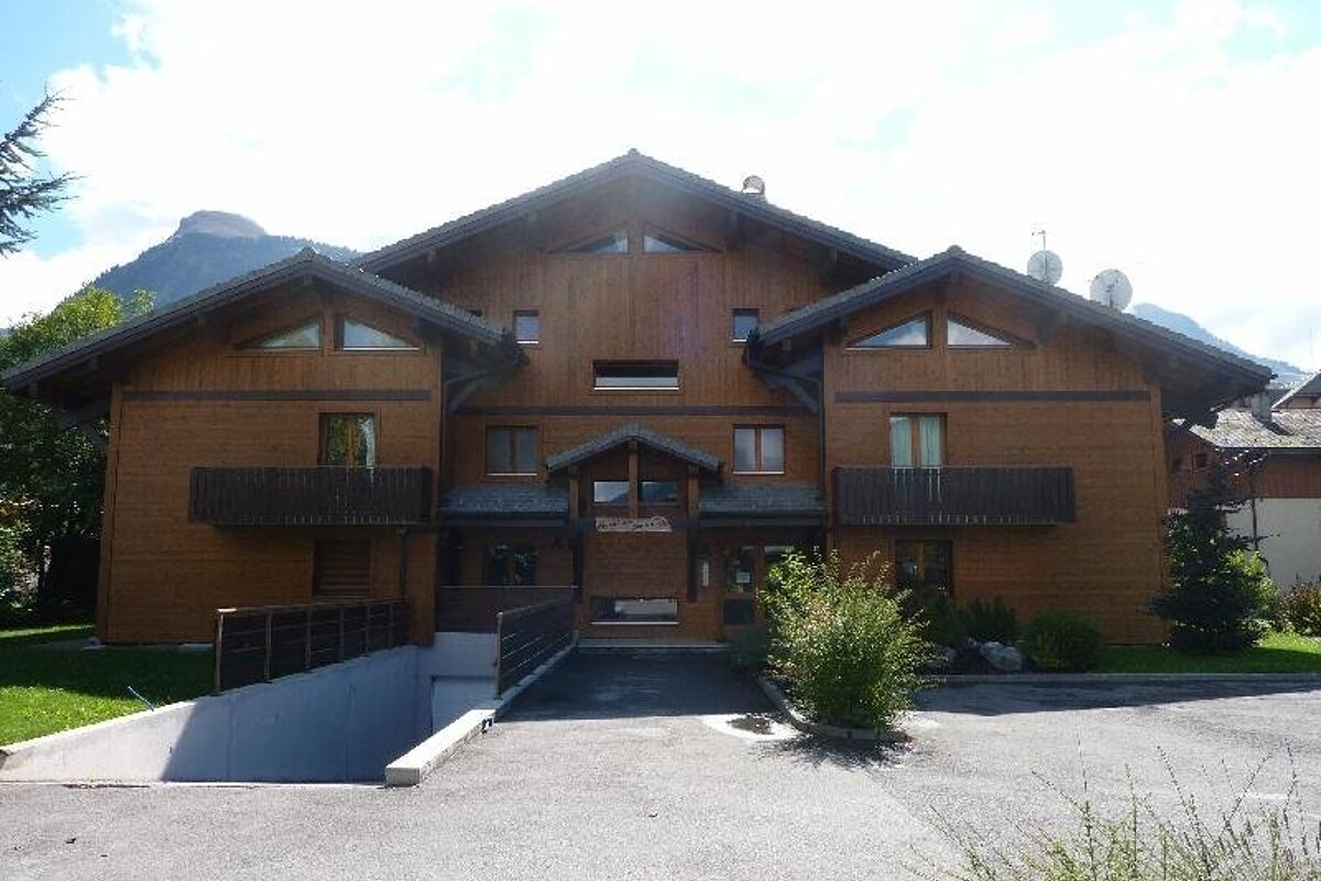 A large wooden chalet-style building with balconies, a garage ramp, and mountains in the background.