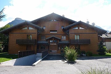 A large wooden chalet-style building with balconies, a garage ramp, and mountains in the background.