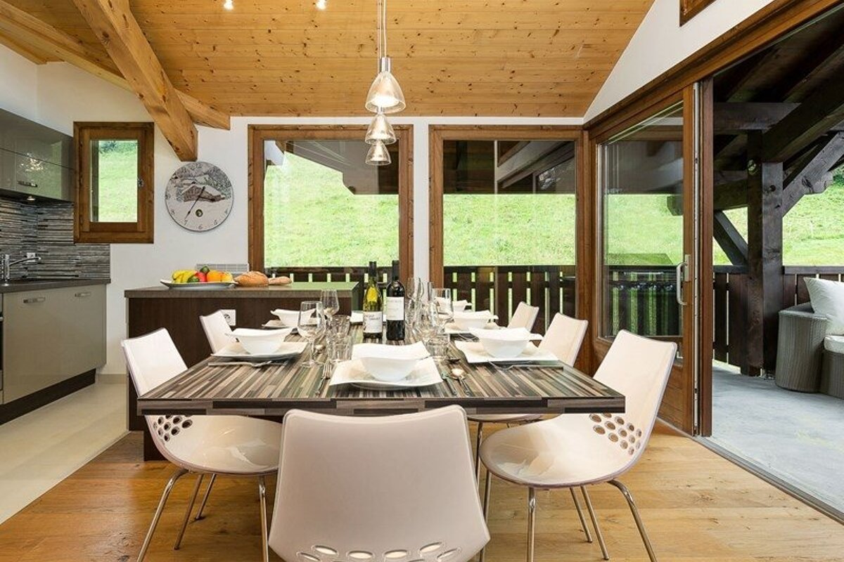 Modern chalet dining room featuring a set table, white chairs, and large windows overlooking a green hillside.