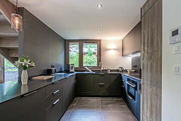 A modern U-shaped kitchen with sleek black cabinets, a tiled floor, wood accent wall, and a window overlooking green trees.