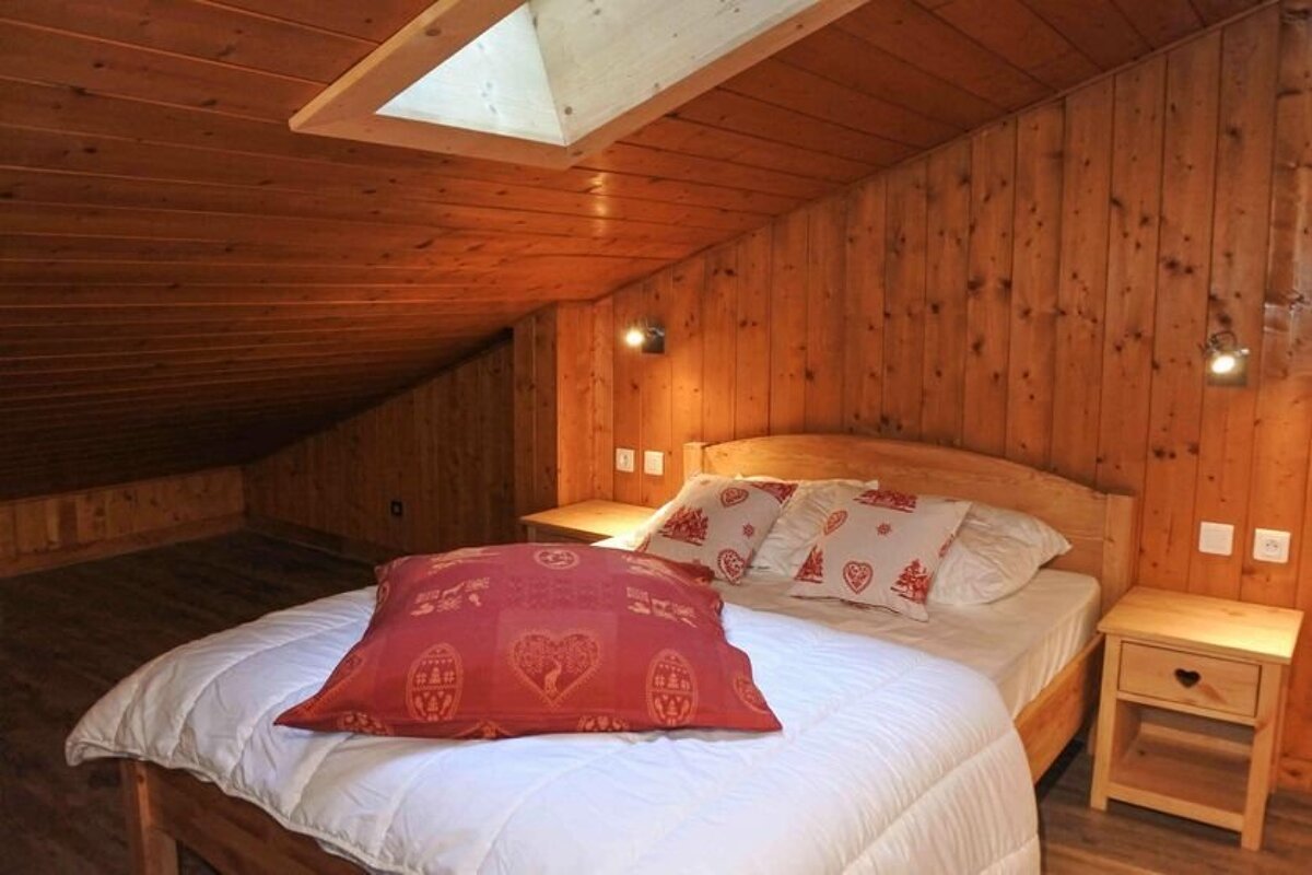 A cozy, wood-paneled attic bedroom with a bed featuring a white duvet and red/white patterned pillows, bedside tables, and a skylight.