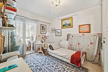 A cozy, eclectic room with a single white metal bed, patterned rug, chandelier, framed art, a sheer-curtained balcony, and cluttered shelves.