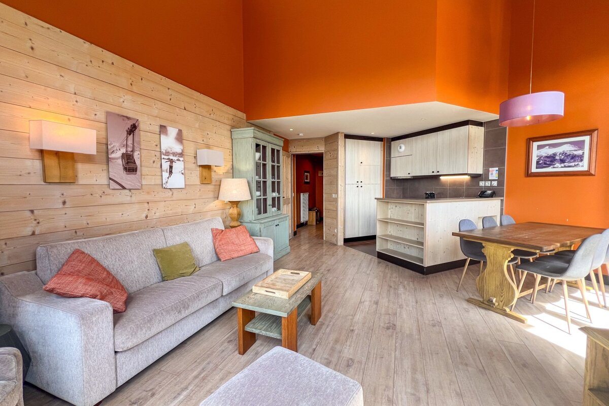 Cozy, modern apartment with a wooden-panel wall, bright orange ceiling, grey sofa, wooden dining set, and open kitchen.