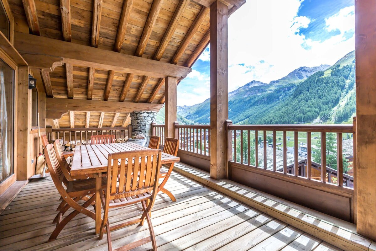 A wooden chalet balcony with a dining table and chairs, offering a stunning view of alpine mountains.
