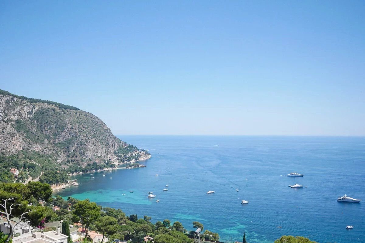 A high-angle view of a vast blue sea with yachts near a green, rocky coastline under a clear sky.