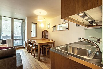 Cozy apartment interior featuring a kitchen sink in the foreground, with a wooden dining table and chairs in the background.