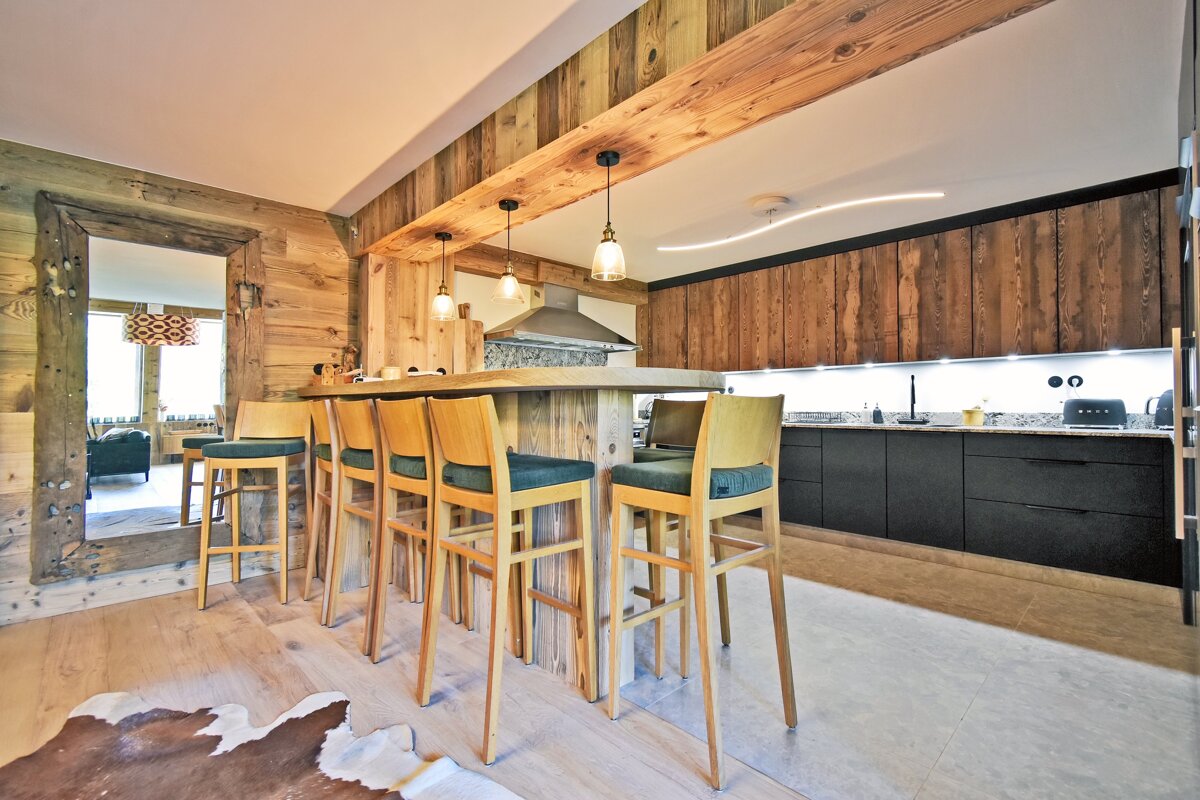 Rustic kitchen featuring a wooden bar with stools, modern black cabinets, and warm wood-paneled walls and ceiling beams.