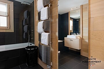 A sleek, modern bathroom featuring dark tiles, light wood, a bathtub/shower, towel warmer with towels, and a floating vanity.