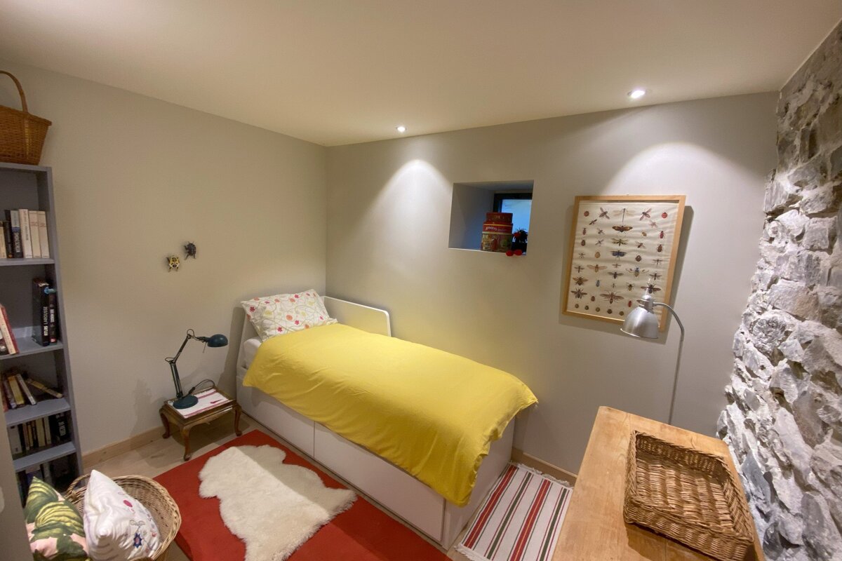 A cozy bedroom features a yellow-duvet bed, bookshelf, insect art, and a rustic stone accent wall, illuminated by warm recessed lights.