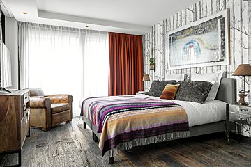 Cozy bedroom featuring birch tree wallpaper, a vibrant striped blanket, rustic wood floor, leather armchair, and large windows with orange/white curtains.