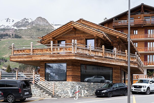 A wooden alpine chalet with large windows and a stone foundation, set against a backdrop of snow-capped mountains.