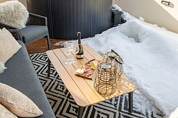 Cozy winter deck with a hot tub, snow, and a wooden table set with champagne, a charcuterie board, and lanterns.